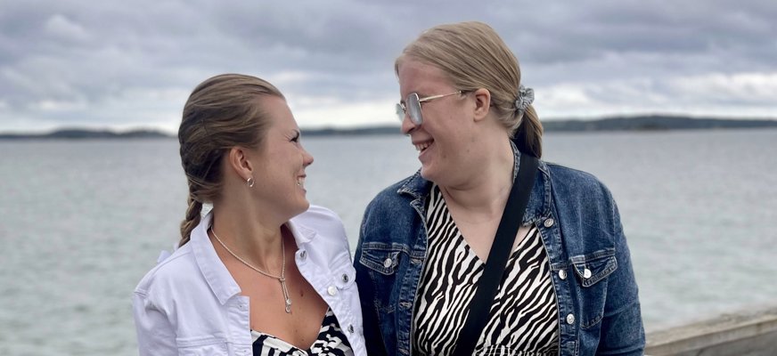 Emelie Pålsson och Rebecka Olsson står och tittar på varandra. De bär matchande svartvita djurmönstrade klänningar och ler. I bakgrunden syns havet.