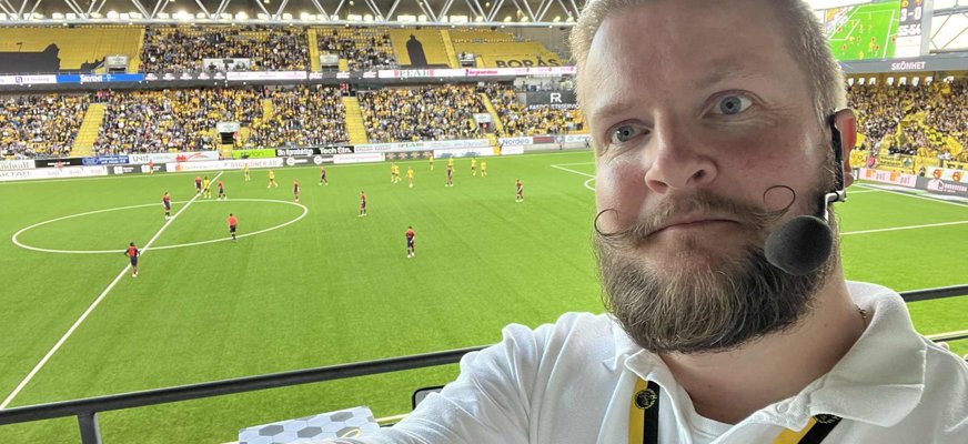 Syntolken Kristoffer Lidén har skägg och mustasch och bär en hörsnäcka med mikrofon. I bakgrunden syns en pågående match på en fotbollsplan.