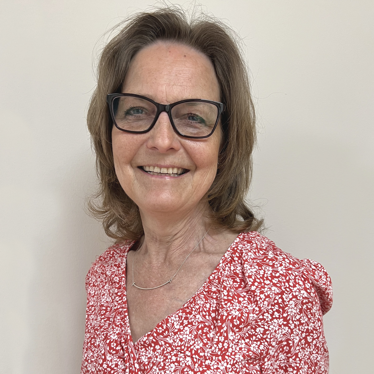 Leende kvinna i 50-årsåldern, brunt axellångt hår, mörka glasögon, silverhalsband och småblommig röd blus.
