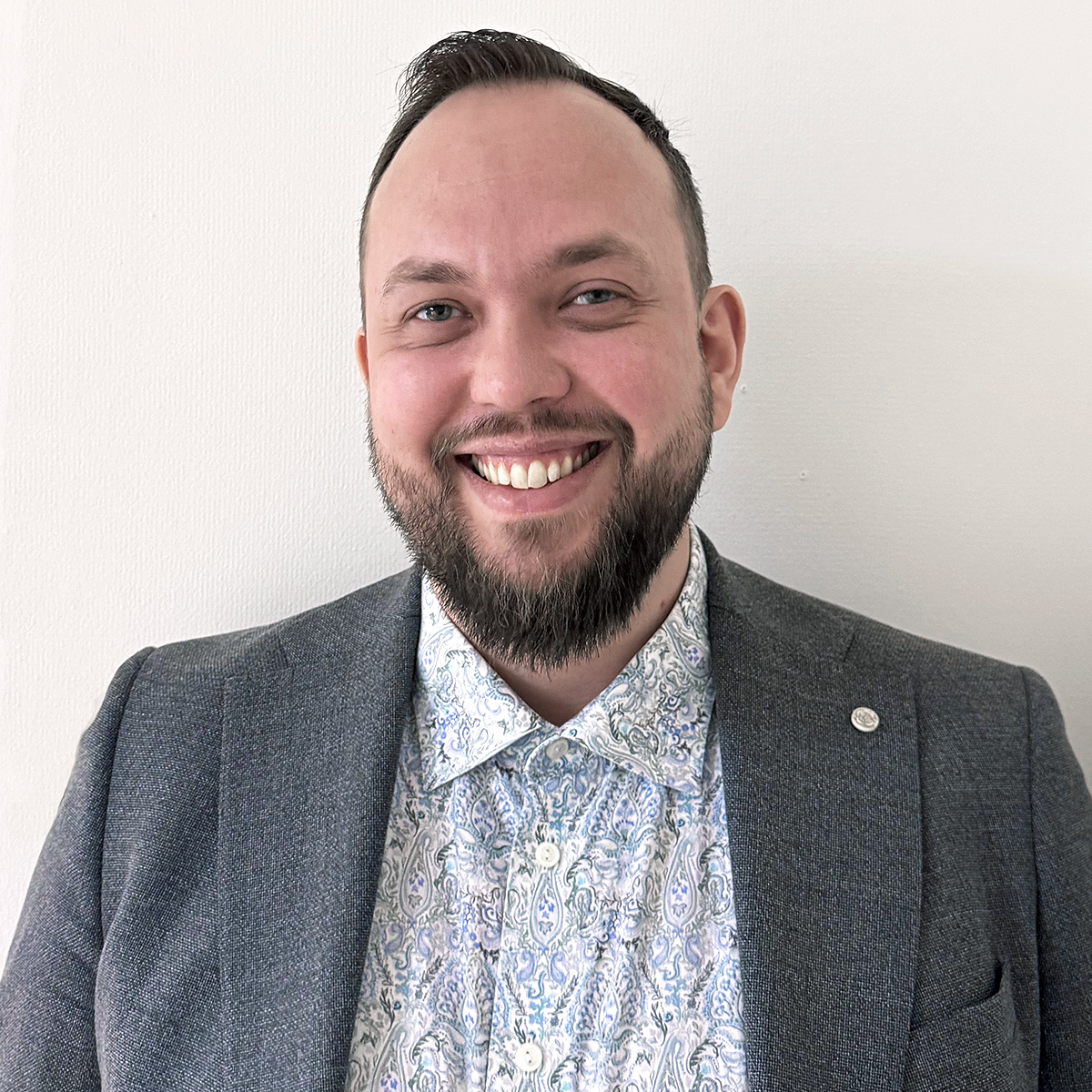 Porträttbild på Anders Ziethén. Anders har kortklippt mörkt hår och kort helskägg. Han har blå ögon och ler stort med tänderna mot kameran. Anders är klädd i en paisleymönstrad helknäppt skjorta med en grå kavaj över.