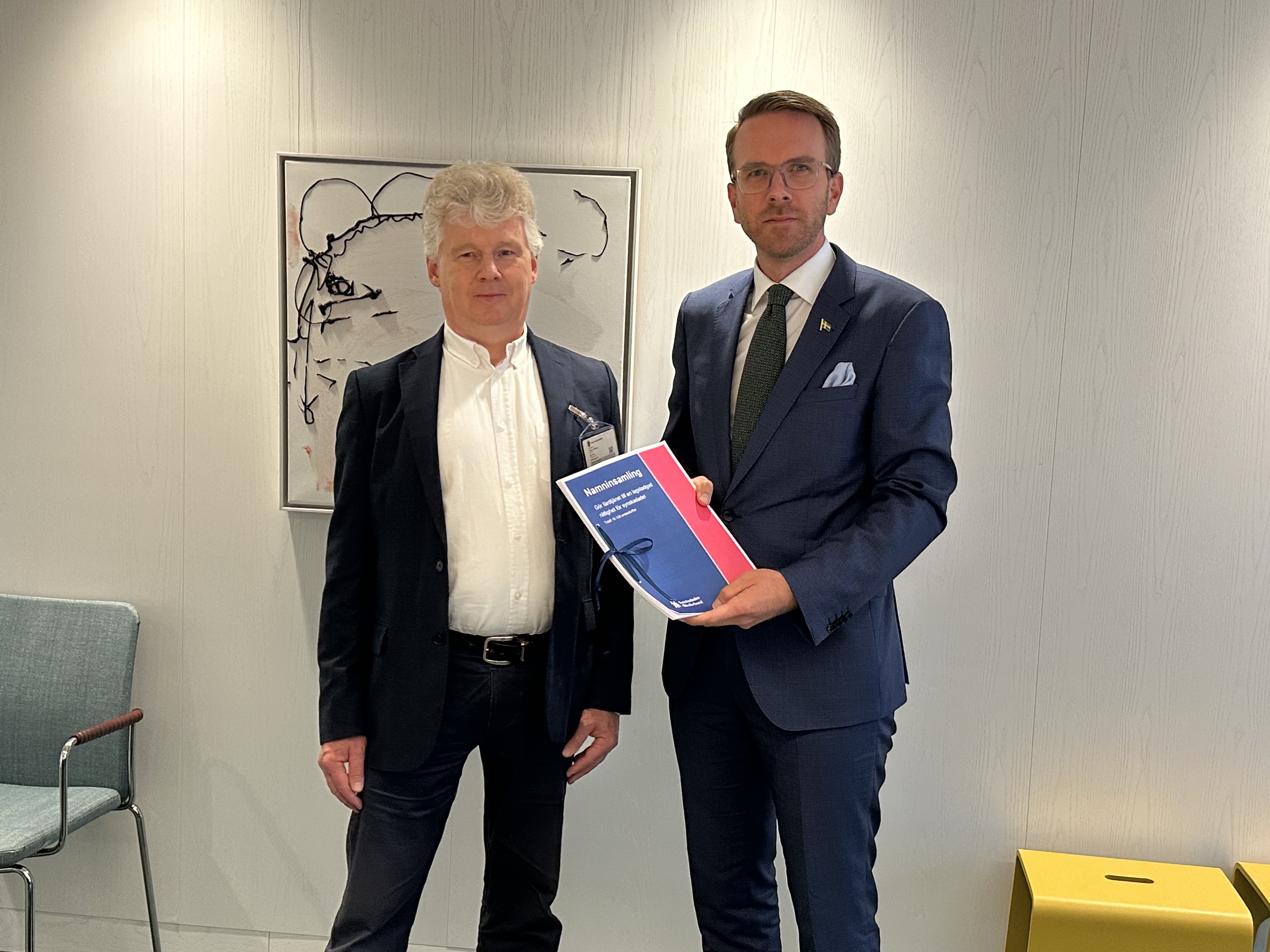 Niklas Mattsson lämnar över namninsamlingen till Andreas Carlson. Båda männen har mörka kavajer och vit skjorta och befinner sig i ett mötesrum på regeringskansliet. Bakom dem skymtar en tavla med ett svartvitt mönster. 