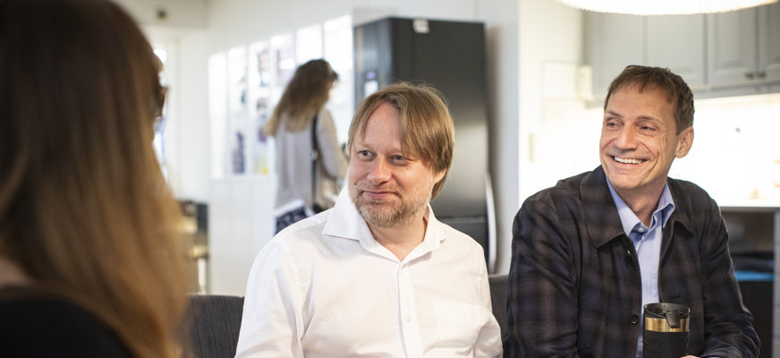 Tre personer sitter tillsammans vid ett bord. De dricker kaffe och pratar leende med varandra.