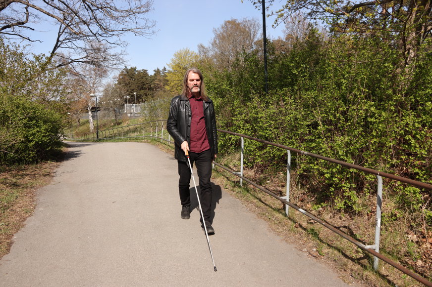 Fredrik går längs en gångväg med sin vita käpp. Han är klädd i röd skjorta, svarta jeans och en svart lång skinnjacka. Han har axellångt brunt hår och mustasch.