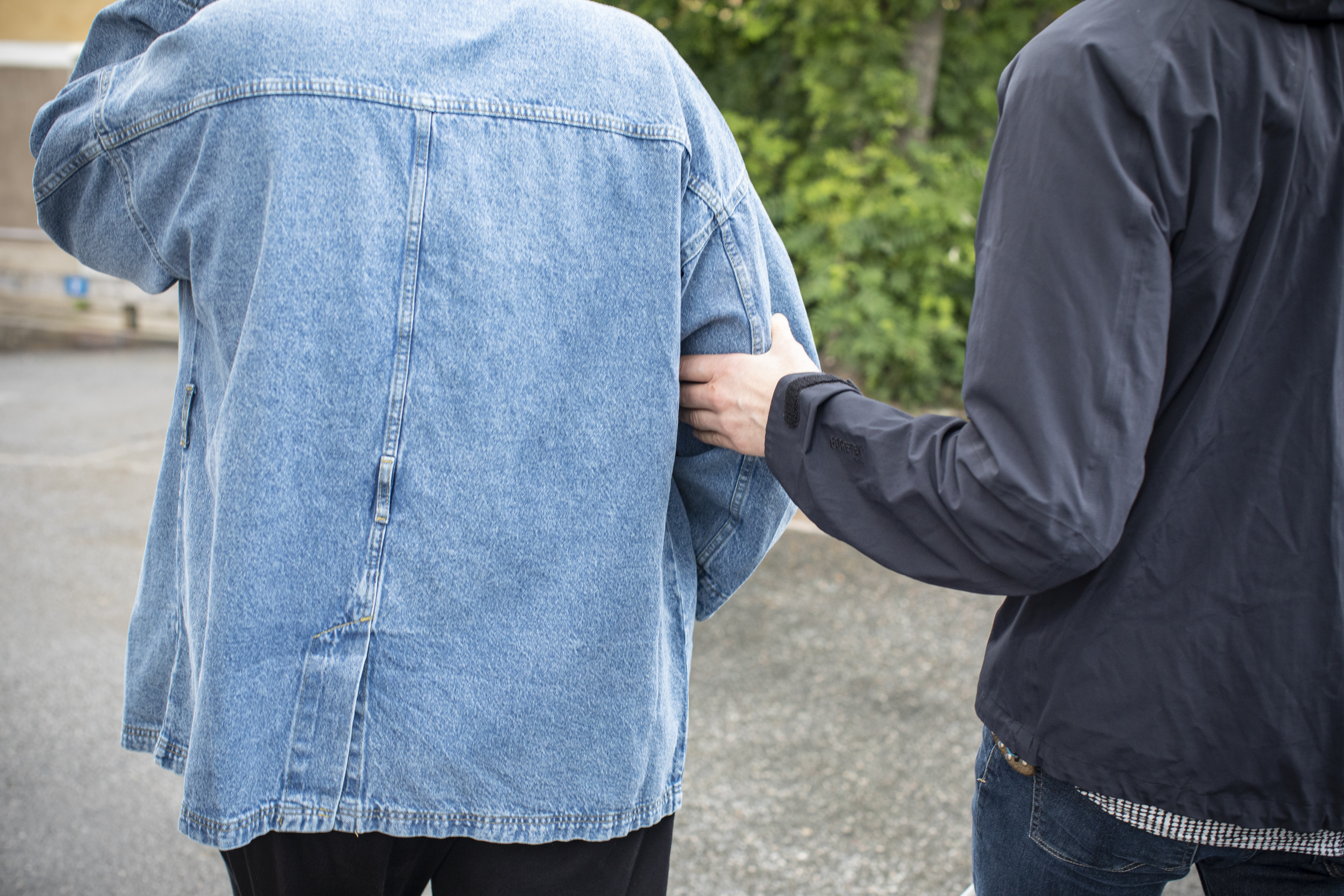 Ryggtavlorna på två personer. Den ena ledsagar den andra. I bakgrunden syns gröna löv.