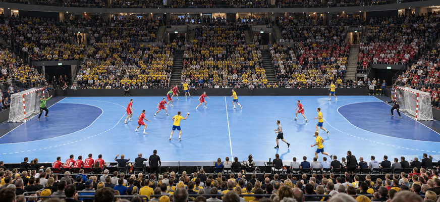 En handbollsmatch sedd från en bit upp på läktaren vid mittlinjen. Sverige spelar i gula tröjor och Österrike i röda, inför fullsatta läktare runt den blå spelplanen.