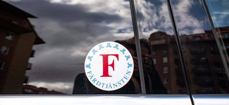 En närbild av en bilruta med ett runt färdtjänstmärke med ett stort rött F. I den blanka rutan speglas moln och byggnader i bakgrunden.