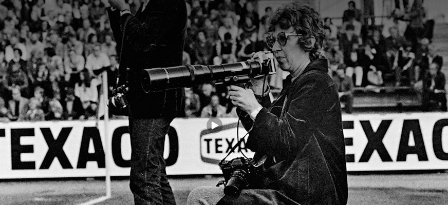Svartvitt fotografi av en kvinnlig fotograf med glasögon och kort krulligt hår som sitter vid sidan av en idrottsarena och fotograferar med ett långt teleobjektiv. En andra kamera med ett standardobjektiv hänger runt halsen och publiken syns i bakgrunden.