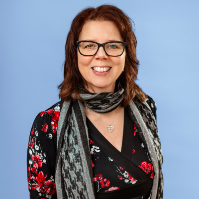 En leende kvinna med axellångt mörkt hår och med glasögon, iklädd en svart långärmad tröja med röda blommönster på. Hon har ett silverfärgat halsband. Bakgrunden går i en ljusblå ton.