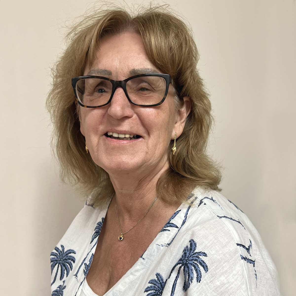 Leende kvinna i 50-årsåldern, axellångt brunt hår med självfall, glasögon med mörk skalm, blåvit mönstrad blus, guldörhängen.