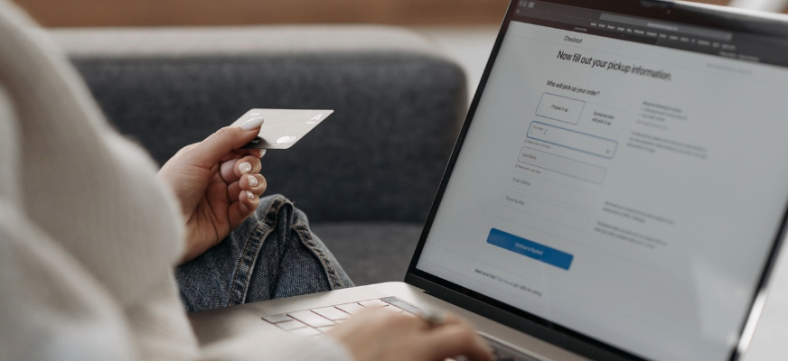En kvinna sitter med datorn i knäet och ett kontokort i ena handen. Hon fingrar samtidigt på datortangenterna och på skärmen syns ett betalformulär.