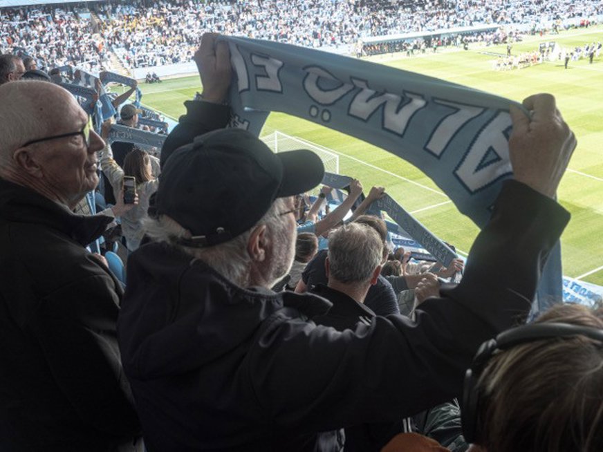 En av gruppens deltagare syns bakifrån med en MFF halsduk som hålls ovanför huvudet och fotbollsplanen syns i bakgrunden.