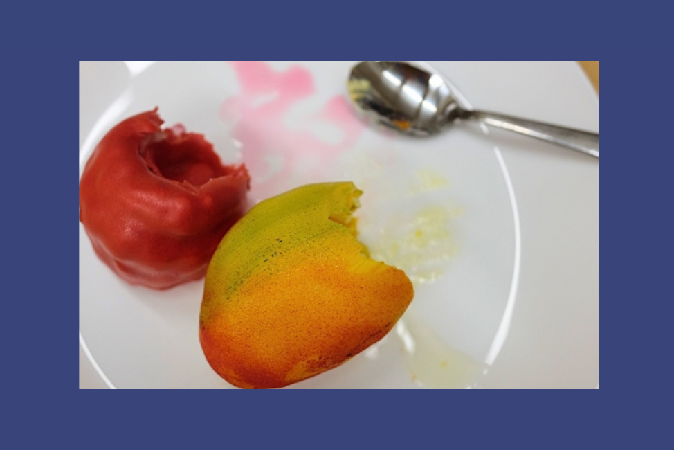 På en vit tallrik ligger två bakelser som ser ut som en gul mango och ett rosa stort hallon.