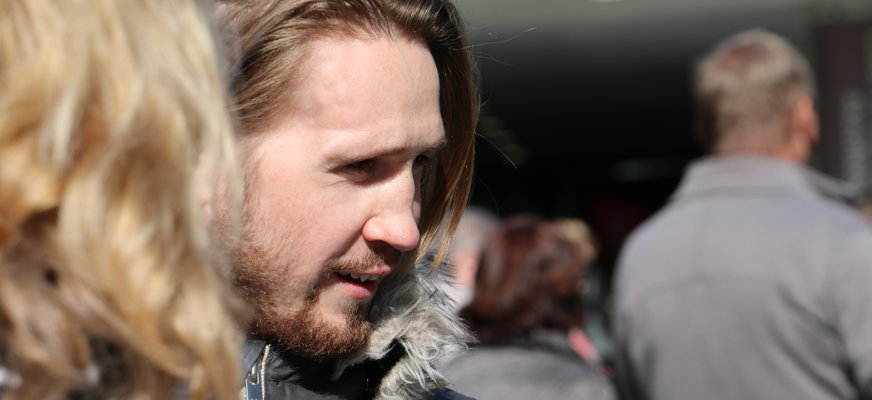 Skäggig man i profil som står i folksamling.