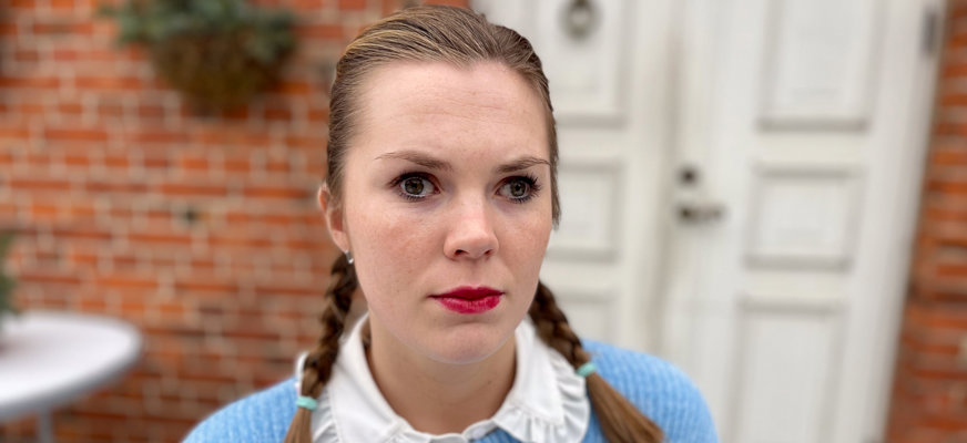 Porträttbild på Emelie som sitter utanför ett rött tegelhus med vit dörr. Emelie har bruna ögon, fräknar och ljusbrunt hår med ljusa slingor uppsatt i två flätor som faller över axlarna. Hon är klädd i ljusblå tröja över en vit blus med spetskrage och har målat läpparna med rött läppstift. Hon ser med allvarlig min in i kameran.