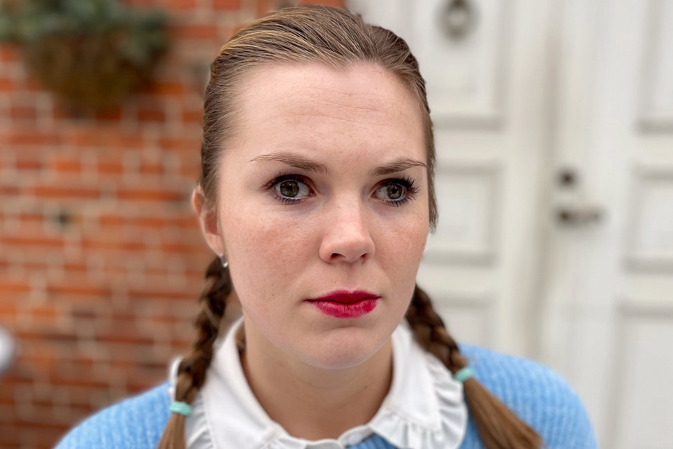 Porträttbild på Emelie som sitter utanför ett rött tegelhus med vit dörr. Emelie har bruna ögon, fräknar och ljusbrunt hår med ljusa slingor uppsatt i två flätor som faller över axlarna. Hon är klädd i ljusblå tröja över en vit blus med spetskrage och har målat läpparna med rött läppstift. Hon ser med allvarlig min in i kameran.