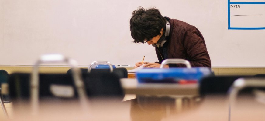 En man med mörkt hår och hörlurar runt halsen sitter ensam i ett klassrum. Han tittar ner på ett papper och har en penna i handen. Mannen på bilden är inte David Rosander.