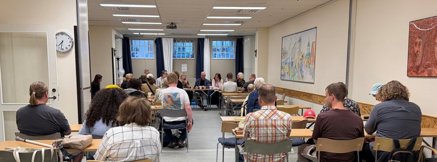 Konferensrum med ett tjugotal åhörare med ryggarna vända mot kameran. Längst fram i rummet en grupp personer som leder mötet. i rummet finns tre fönster med gardiner, en klocka samt en färgglad tavla på väggen