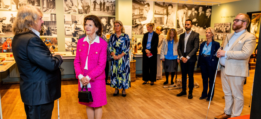 Bild från Synskadades Museum. På väggarna syns fotografier. Drottning Silvia står i mitten i en rosa dräkt och håller en svart handväska. Till vänster står Håkan Thomsson och håller i sin vita käpp. Han är långhårig och bär mörk kostym. Till höger står Linus Johansson i ljus kostym med vit käpp. Bakom dem står flera personer. En ledarhund ligger på golvet.