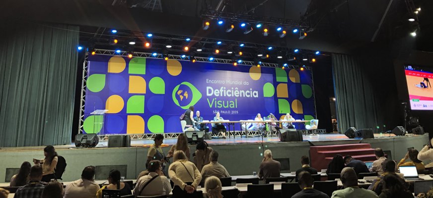 En scen med en grupp människor som sitter uppradade vid ett bord. I bakgrunden en stor skärm med texten "Encontro Mundial da Deficiencia Visual - Sao Paulo 2025". Framför scenen sitter människor och lyssnar.