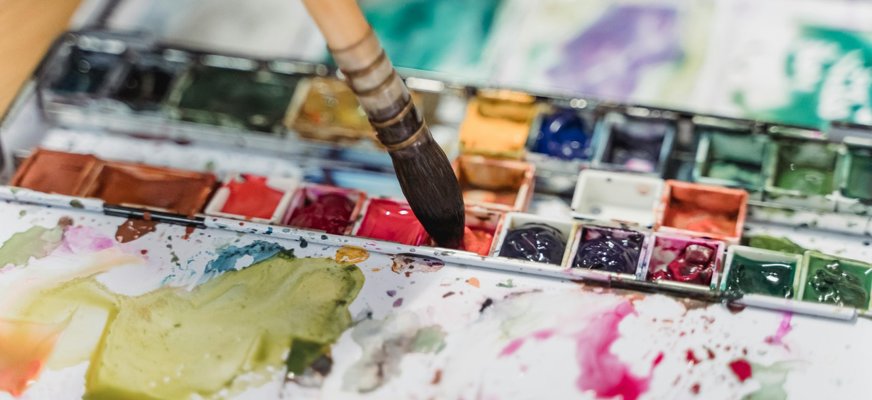 En närbild på en pensel. Den doppas i en av färgerna i ett etui med akvarellfärger.