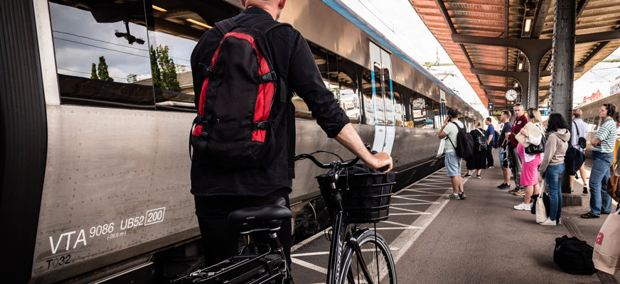 En man med keps och ryggsäck leder en cykel på en tågperrong. Till vänster syns ett tåg och längre bort syns många resenärer.