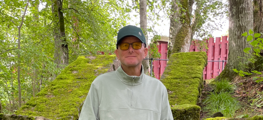 En man med keps och tonade glasögon står utomhus framför mossbeklädda stenmurar. Han bär en ljus tröja med hög krage och tittar mot kameran. Runt omkring syns träd och grönska, och i bakgrunden skymtar ett rött staket.
