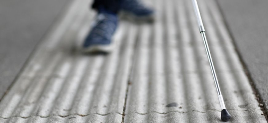 En person som använder vit käpp går långs ett ledstråk. Enbart skorna, delar av de mörka byxorna och nedre delen av käppen syns samt det vita ledstråket som kontrasterar mot den mörkare gatan.