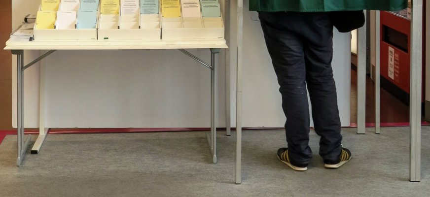 Två ben sticker ut under ett skynke i ett röstningsbås i en vallokal. Bredvid båset syns blå, vita och gula valsedlar prydligt uppradade.