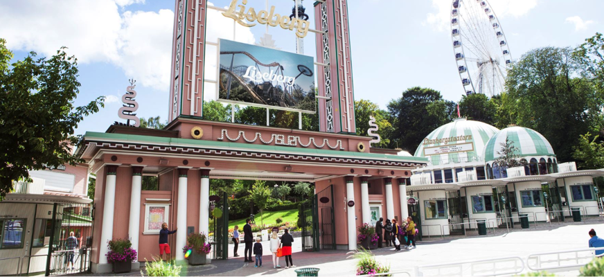 ngången till nöjesparken Liseberg i Göteborg, med den klassiska entréportalen, Lisebergsteatern till höger och det stora pariserhjulet i bakgrunden. Några besökare syns vid entrén under en solig dag.