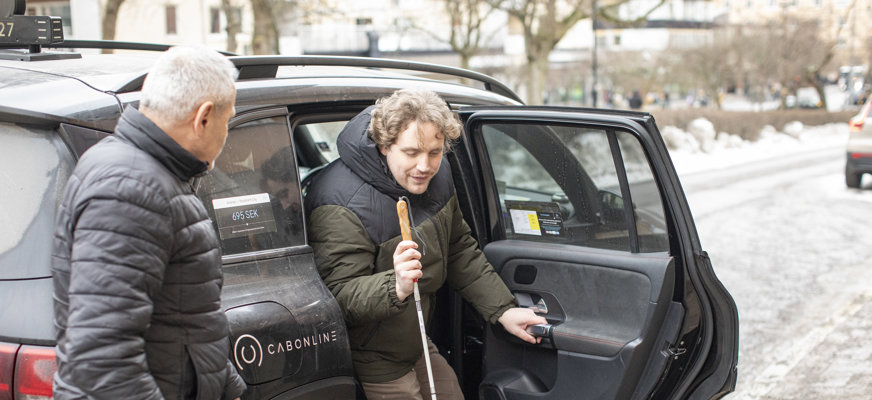 En man med vit käpp stiger ut ur en svart färdtjänstbil. Chauffören står redo att assistera mannen.