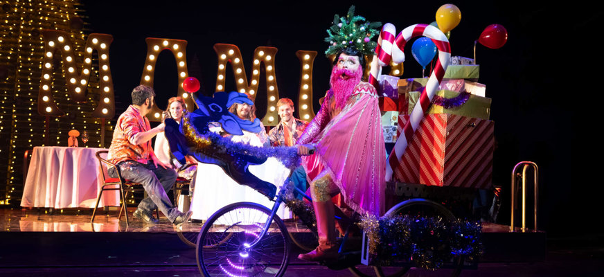 En färgstark scenföreställning där en person i glittrig dräkt cyklar förbi med presenter medan andra artister sitter vid ett bord på scenen.
