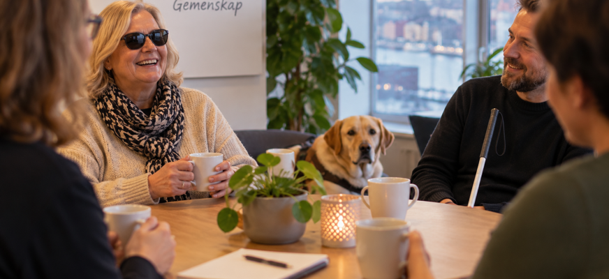 En mindre grupp personer samtalar runt ett bord med kaffe. En vit käpp och en ledarhund syns i rummet, som signalerar gemenskap och erfarenhetsutbyte kring livet med synnedsättning.