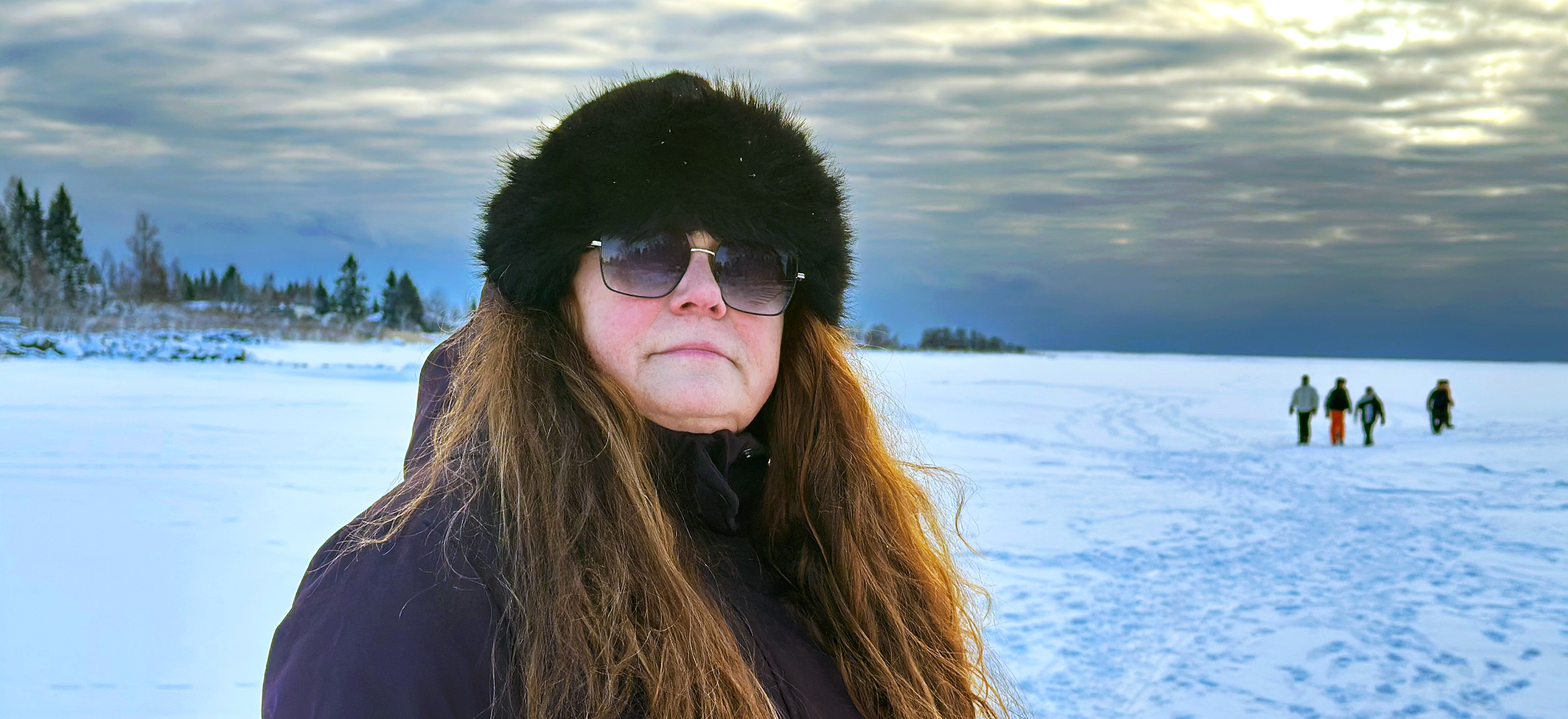 Porträttbild på Ann som står på isen, på ett fruset vattendrag, i solnedgången. I bakgrunden skymtar fotspår i snön och en grupp människor på isen. Ann har rödskiftande långt hår och är röd om kinderna. Hon bär en mörk jacka, pälsmössa och solglasögon.