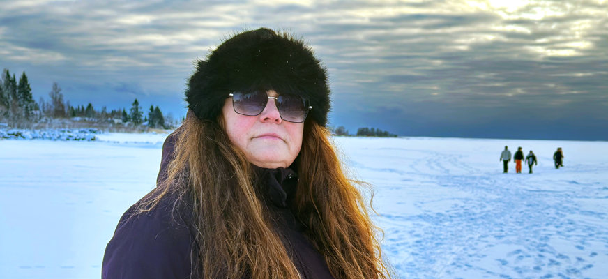 Porträttbild på Ann som står på isen, på ett fruset vattendrag, i solnedgången. I bakgrunden skymtar fotspår i snön och en grupp människor på isen. Ann har rödskiftande långt hår och är röd om kinderna. Hon bär en mörk jacka, pälsmössa och solglasögon.