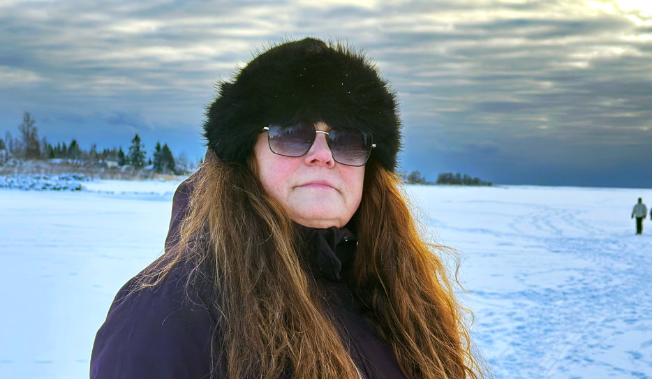 Porträttbild på Ann som står på isen på ett fruset vattendrag, i solnedgången. I bakgrunden skymtar fotspår i snön och en grupp människor på isen. Ann har rödskiftande, vågigt, långt hår och är röd om kinderna. Hon bär en mörk jacka, pälsmössa och solglasögon.