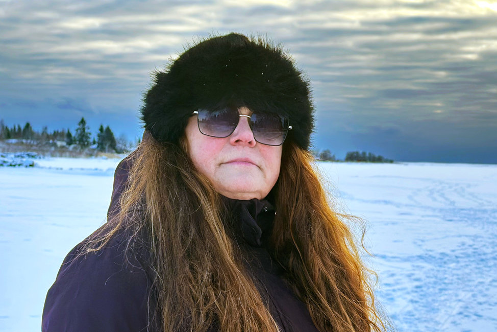 Porträttbild på Ann som står på isen på ett fruset vattendrag, i solnedgången. I bakgrunden skymtar fotspår i snön och en grupp människor på isen. Ann har rödskiftande, vågigt, långt hår och är röd om kinderna. Hon bär en mörk jacka, pälsmössa och solglasögon.