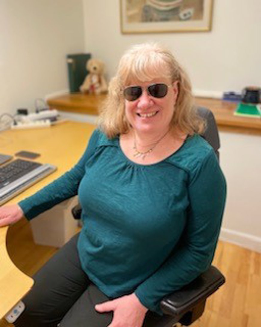 Eva Hildursmark, foto taget på evas rum, eva sitter vid sitt skrivbord har en turkosgrön tröja samt svarta byxor och svarta glasögon.