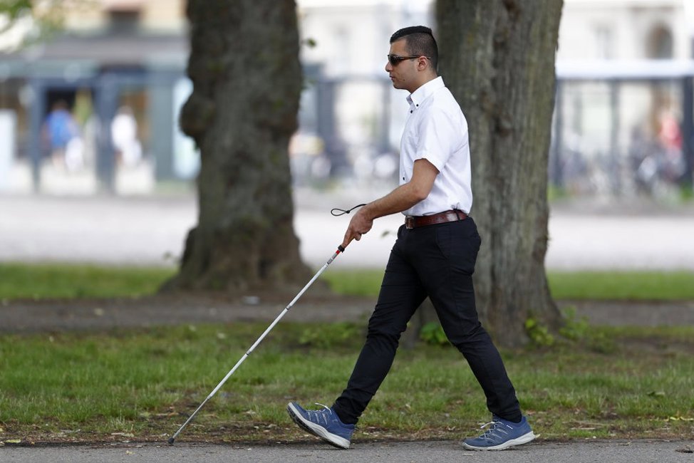 En yngre man med kort mörkt hår och brun hy går med sin vita käpp på trottoar. Han har vit t-tröja och svarta jeans. Vägen ligger vid grönområde med gröna träd. Sommar.