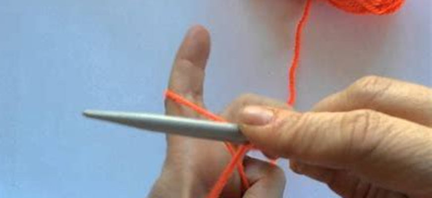 Närbild på två händer som lägger upp maskor på en sticka i en skarp orangeröd nyans.