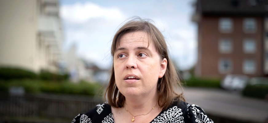 Anna står utomhus framför några lägenhetshus. Hon är en kvinna i 40-årsåldern med kortklippt brunt hår och bruna ögon. Anna bär en svartvitmönstrad skjorta och ett guldhjärta runt halsen. Hon tittar snett uppåt och läpparna är lite särade. 