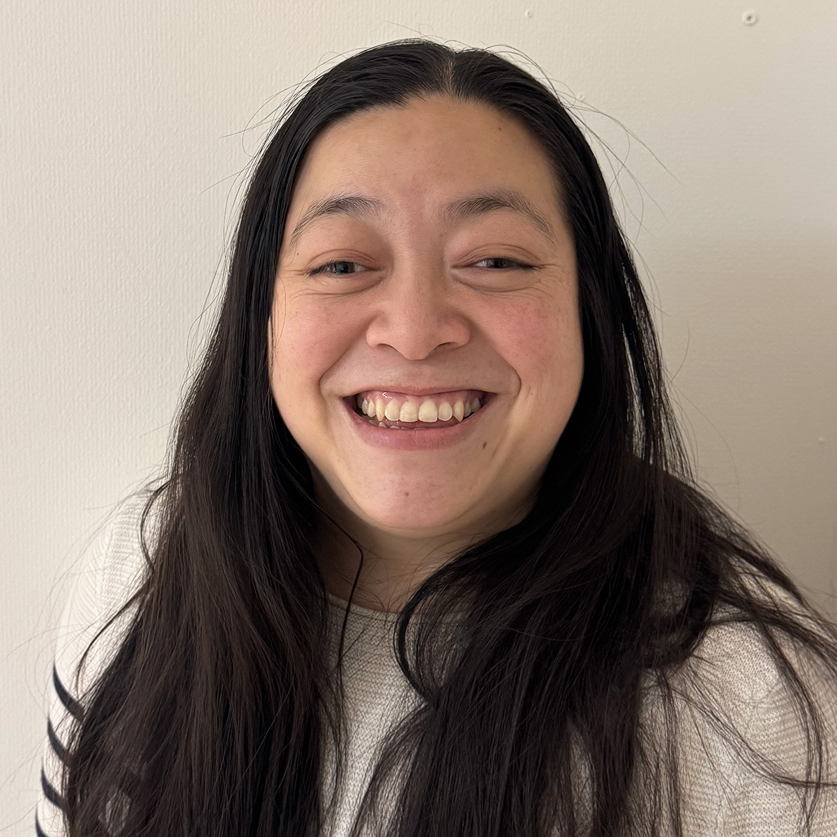 Kvinna med långt rakt och mörkt hår utsläppt över axlarna. Hon har bruna glada ögon ler stort mot kameran. Hon är iklädd en vit tröja med svarta ränder.