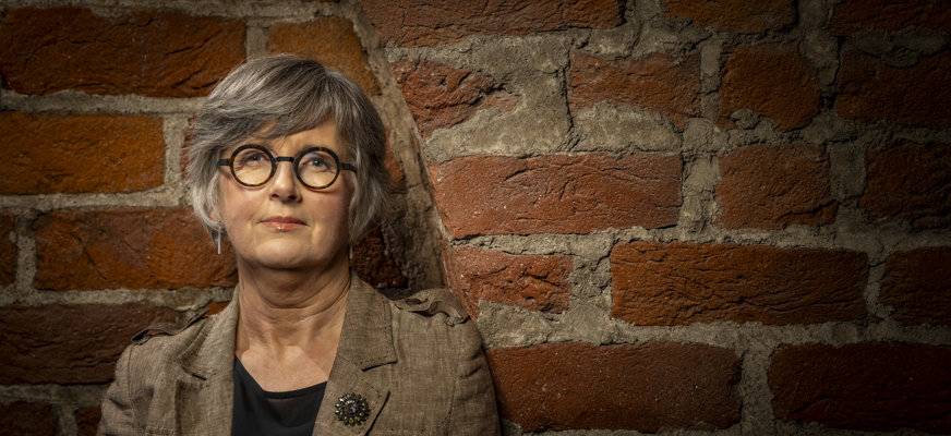 Medelålders kvinna står lutad mot röd tegelvägg. Hon har kort grått hår med snedlugg och bär runda glasögon med tjocka svarta bågar. Hon bär en beige kavaj med svart tröja under.