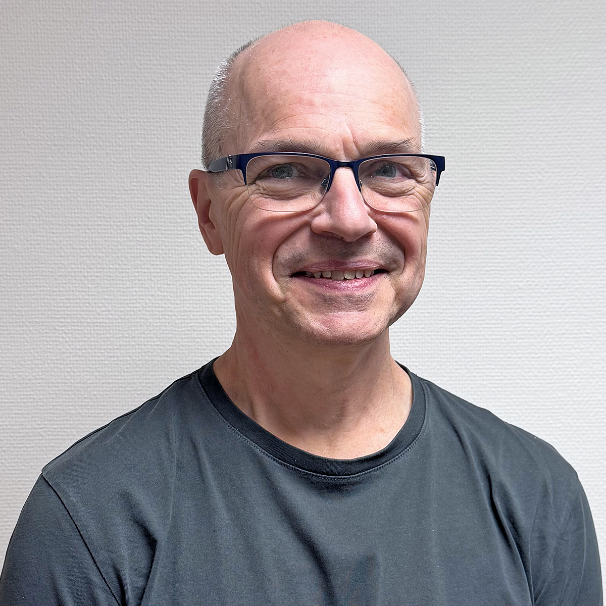Porträttbild på Norbert, en medelålders man. Norbert är blåögd, tunnhårig och renrakad. Han bär glasögon med svarta bågar och en grå t-shirt. Norbert ler mot kameran.