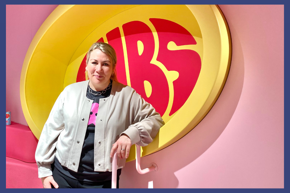 Joanna Mossberg, kommunikationschef BUBS. 35 årig kvinna med blont hår i tofs, beige kofta och svart t-shirt under. Står vid rosa vägg med stor logotype av ”BUBS”- rosa bokstäver i ljusgul oval. Foto: Aziza Dhaouadi
