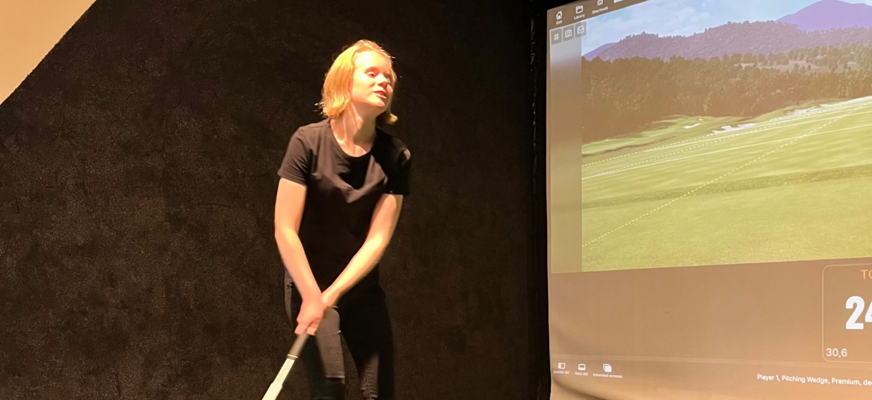 Alicia i svart T-shirt, svarta jeans och mörka sneakers står på en grön konstgräsmatta med en golfklubba i händerna.