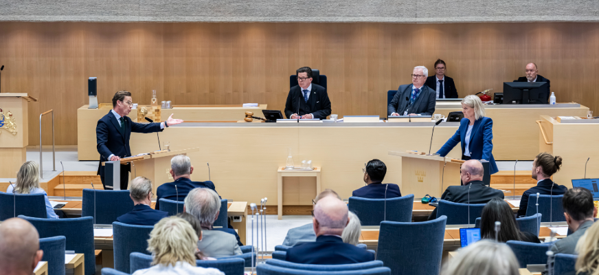 En politisk debatt i en stor sal. Sveriges statsminister Ulf Kristersson står vid talarstolen till vänster och Magdalena Andersson står vid talarstolen till höger. Framför dem sitter åhörare i rader av blå stolar.