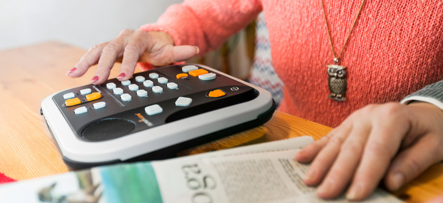 En person sitter och lyssnar på en Daisy-spelare. Bredvid sitter en annan person som läser en papperstidning.