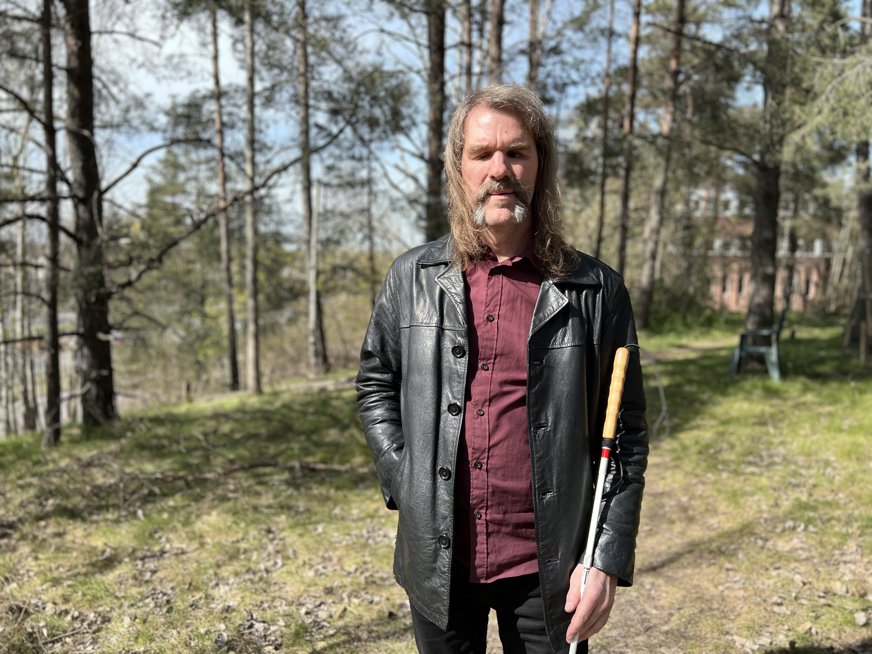 Fredrik står utomhus med träd bakom sig. Han är klädd i röd skjorta, svarta jeans och en svart lång skinnjacka. Han har axellångt brunt hår och mustasch.