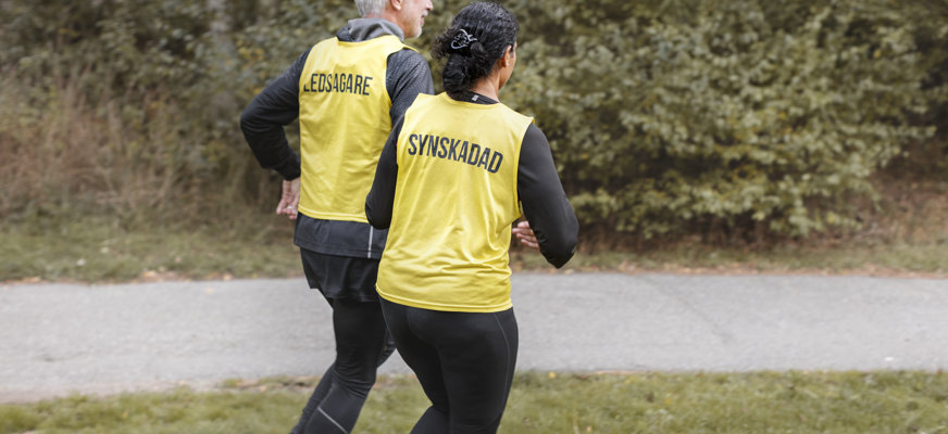 Två personer är ute och springer. De bär båda gula västar, den ena med texten "synskadad", den andra med texten "ledsagare".