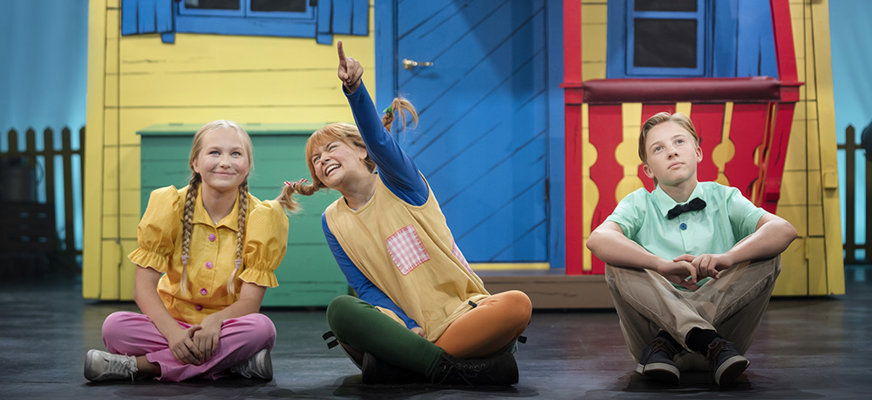 En bild från föreställningen Pippi sitter framför Villa Villerkulla och pekar upp i himlen. Bredvid henne sitter Annika till vänster och Tommy till höger och tittar på det som pippi pekar på.